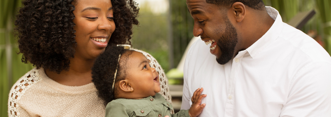 mãe e pai com bebê. Bebê está sorrindo em resposta ao sorriso do pai.