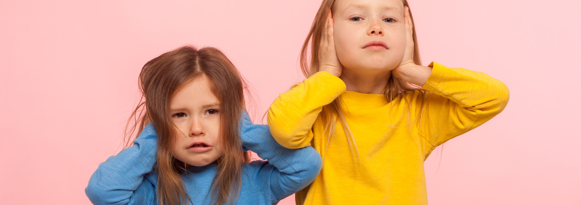 duas meninas, uma de blusa azul e outra de blusa amarela, com as mãos tapando os ouvidos. Fundo rosa.